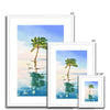 Palm tree set against blue sky and reflecting in clear turquoise water. Fine art print in a white frame with a white mount. Different sizes, smallest at the front.